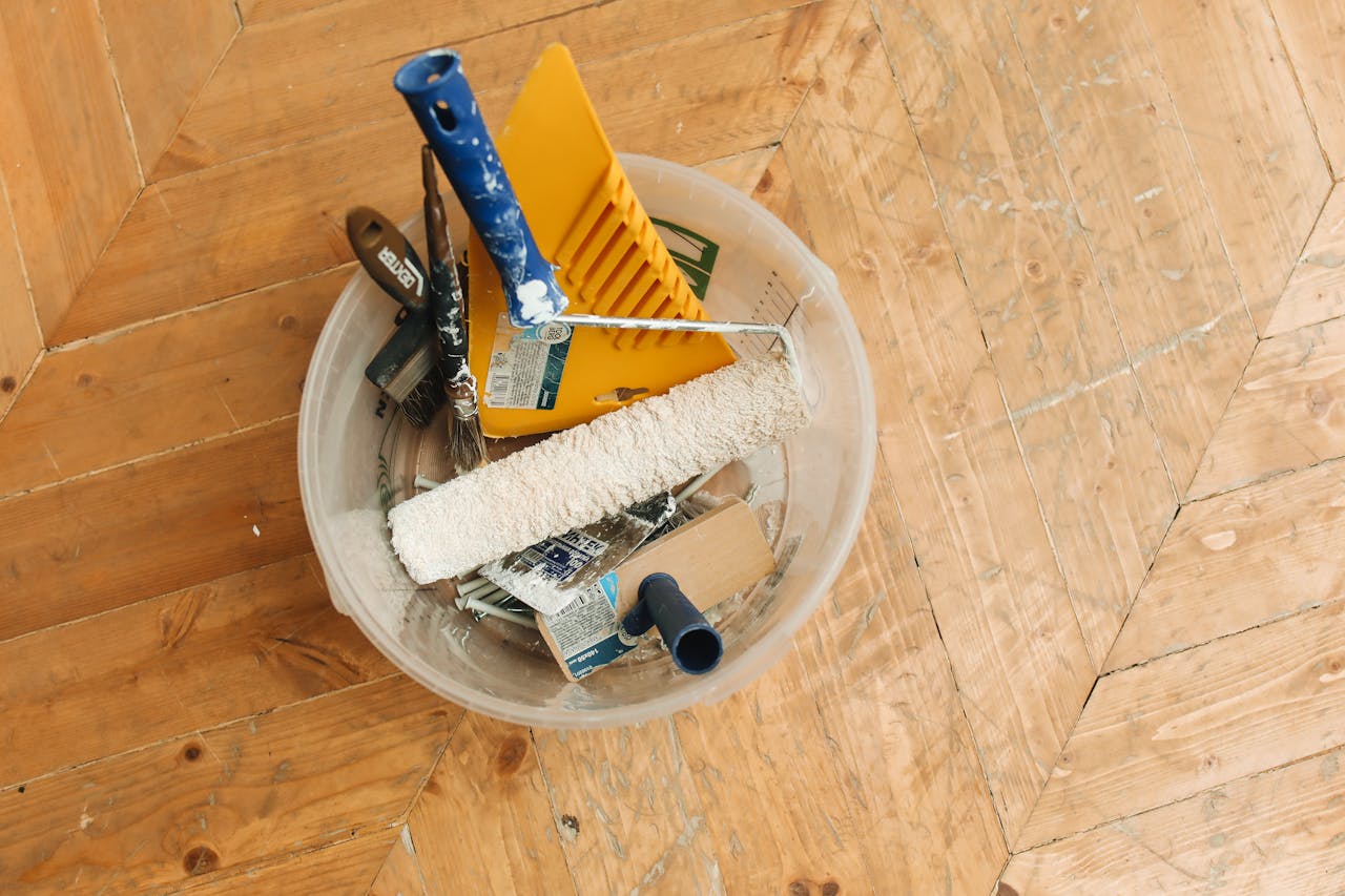 about-03 A collection of home renovation tools in a bucket on a wooden floor, ready for a painting project.